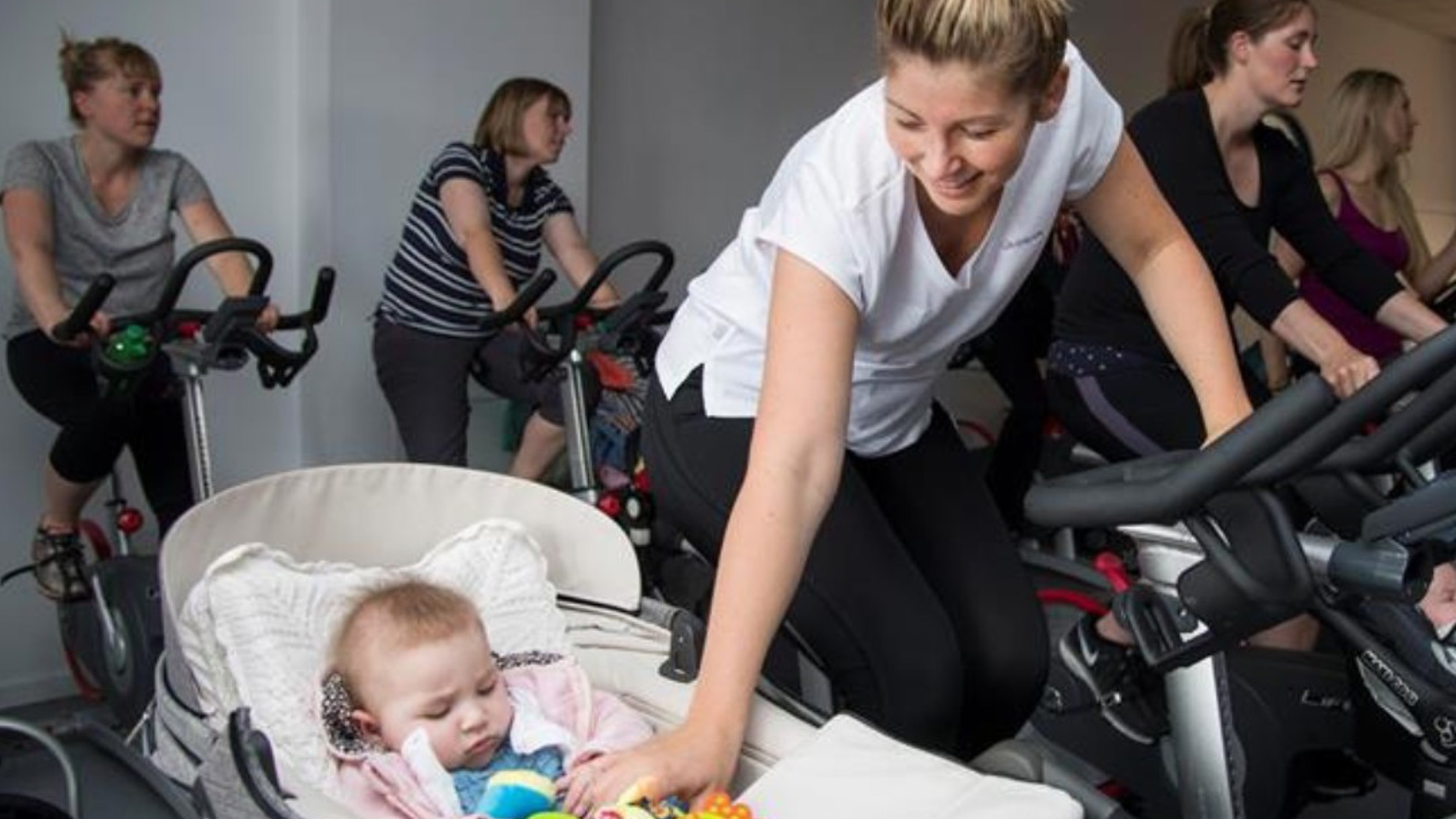 A spin class including one participant reaching down to soothe their baby who is in a pram beside their bike