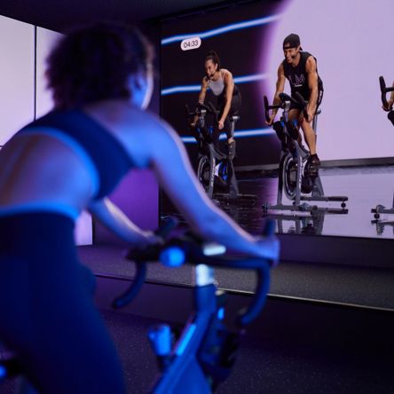 Someone on a spin bike in a dark indoor cycling studio, following an instructor leading the class from a big screen on the wall