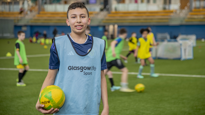 A young person standing facing the camera with a football in their hand as others play football behind them