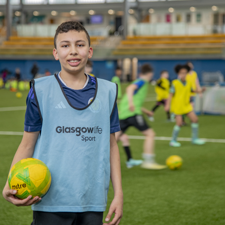 A young person standing facing the camera with a football in their hand as others play football behind them