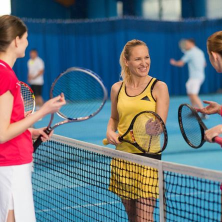 Four women stood two either side of the net on a tennis court, holding racquets and talking 