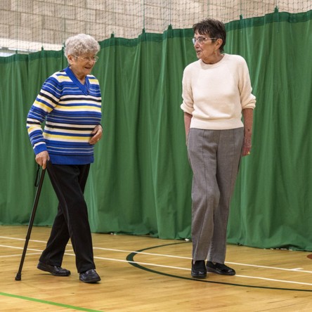 A volunteer guides a smiling older person, who is walking with a stick, between markers laid on a sports hall floor
