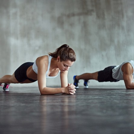 Two people in a plank position on the floor of a grey, modern looking fitness studio