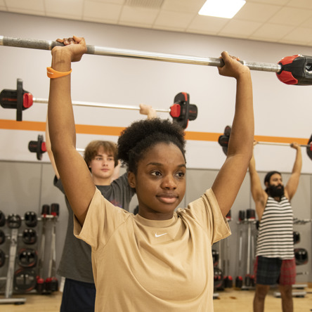 Group fitness class members holding a barbell above their heads with arms straight and locked 