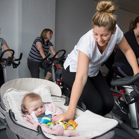 Person on exercise bike with baby in pram beside them playing with a toy 