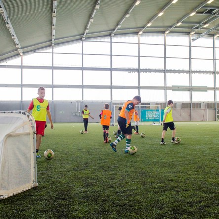A group of children practicing dribbling a football on an indoor pitch