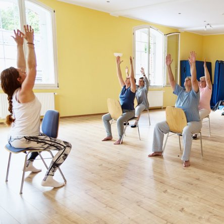A group of people seated on chairs in a fitness studio wiht their arms raised in a Chair Yoga class